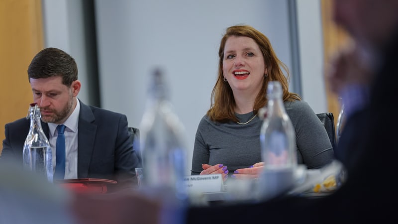 Alison McGovern smiling and speaking at a meeting table with a name card and water bottles in front of her, a man in a suit seated beside her