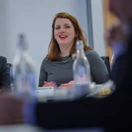 Alison McGovern smiling and speaking at a meeting table with a name card and water bottles in front of her, a man in a suit seated beside her