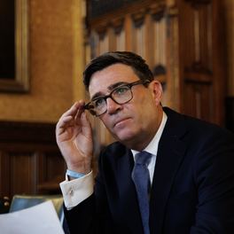 Andy Burnham seated at a table in his Parliamentary Office, adjusting his glasses, with papers in front of him.