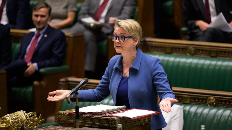 Yvette Cooper speaking at the despatch box in the House of Commons, in a blue jacket and blue-framed glasses, with both hands held out.