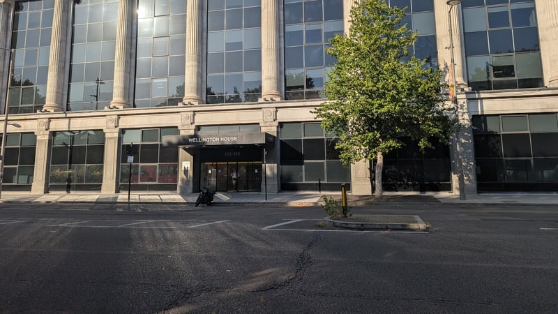 Stone and glass office building with tall columns and a sign reading Wellington House above the entrance, a young tree on the pavement and an empty road in front