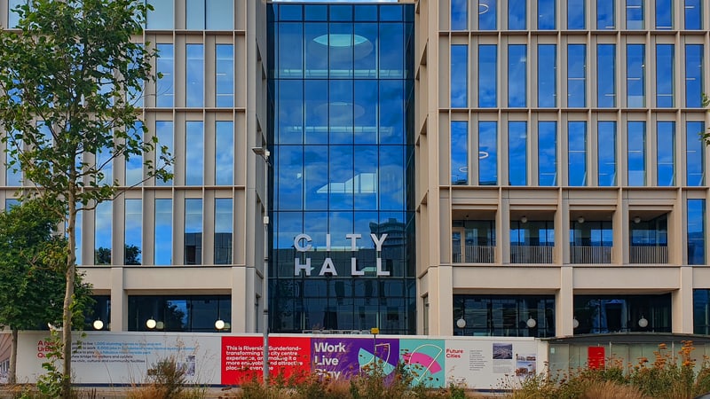 Modern pale stone office building with blue glass windows and a glass central section bearing the words City Hall, with a young tree and hoarding in front