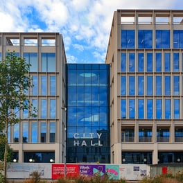 Modern pale stone office building with blue glass windows and a glass central section bearing the words City Hall, with a young tree and hoarding in front